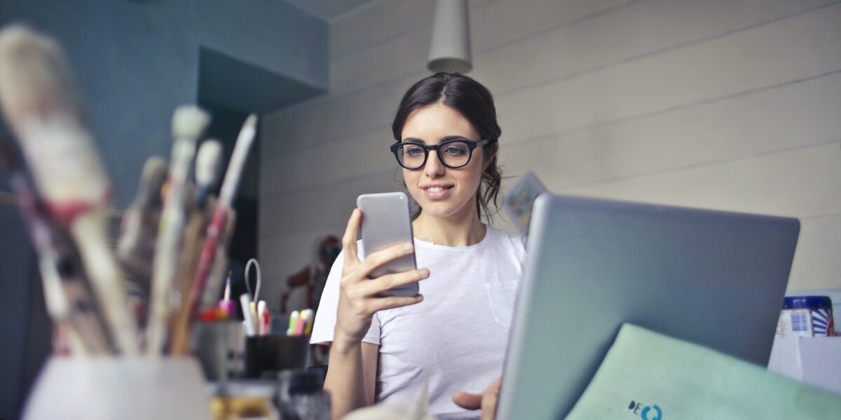 Woman in White T-shirt Holding Smartphone in Front of Laptop